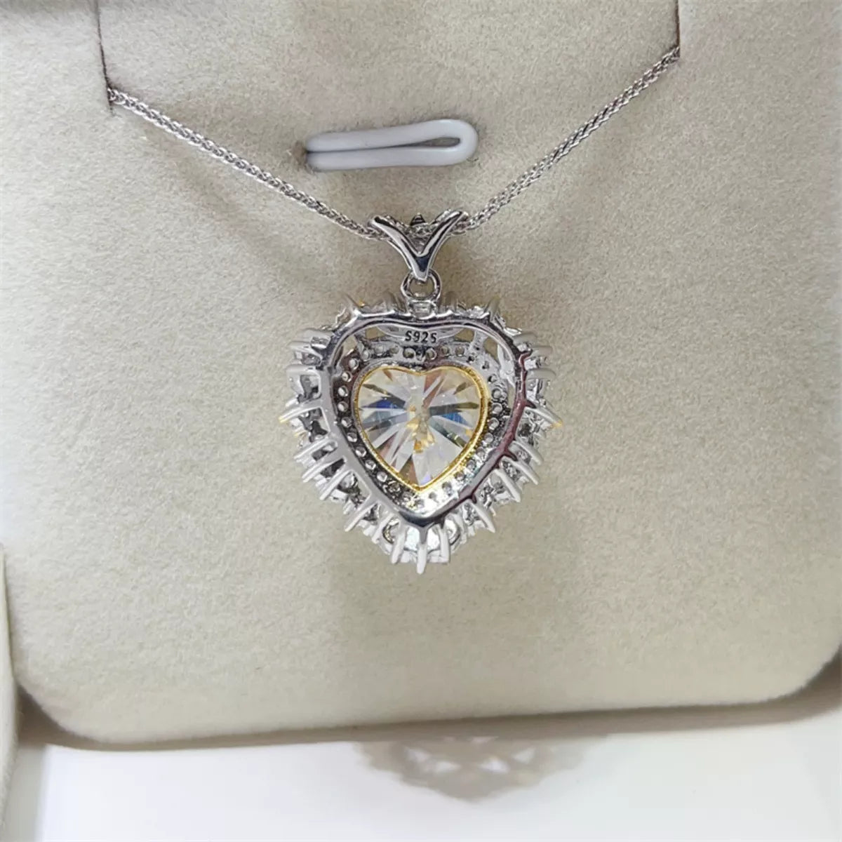 Heart-shaped pendant with a clear center on a silver chain, displayed on a beige cushion.