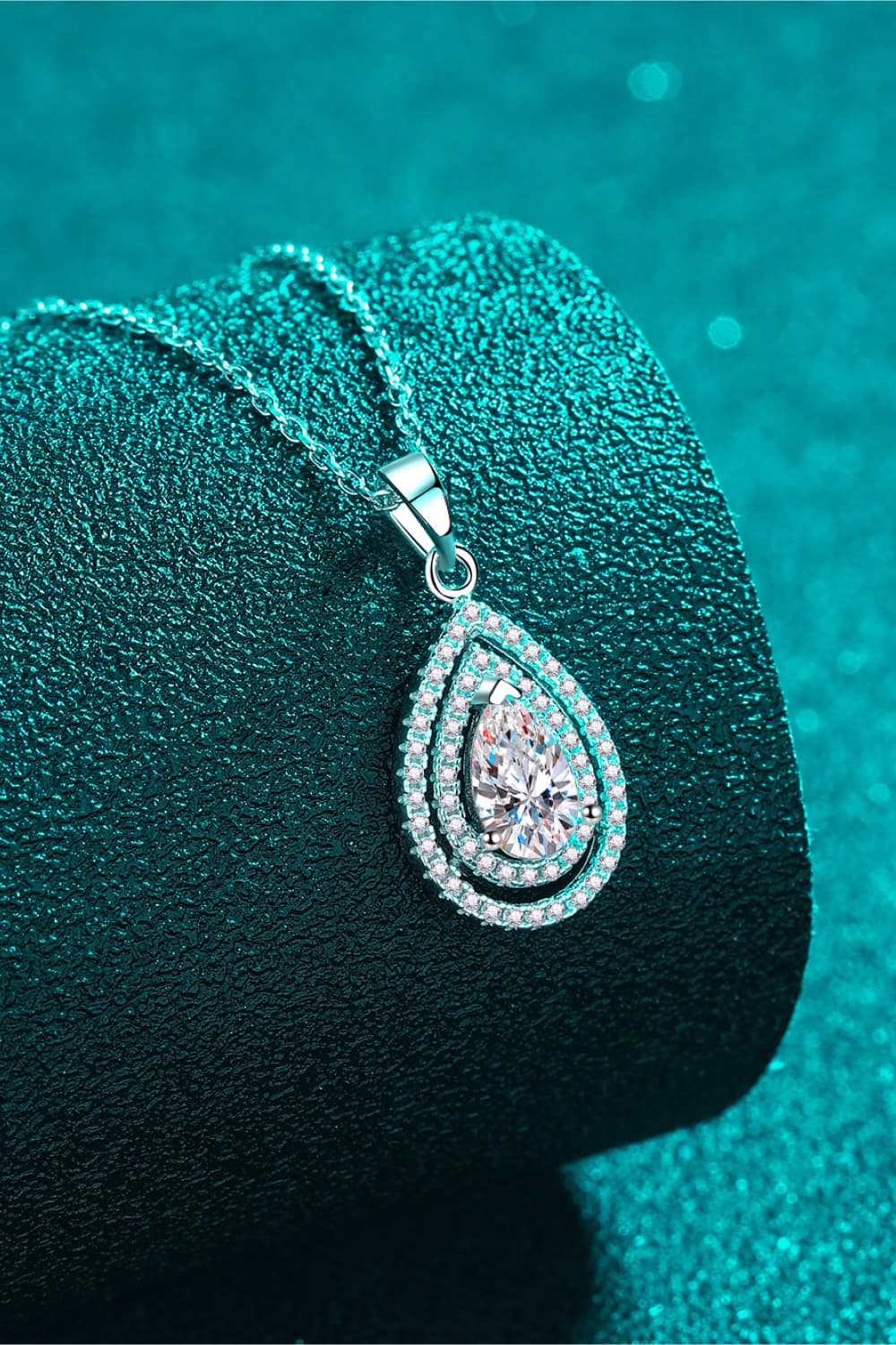 Teardrop-shaped diamond pendant on a black stand with a teal background