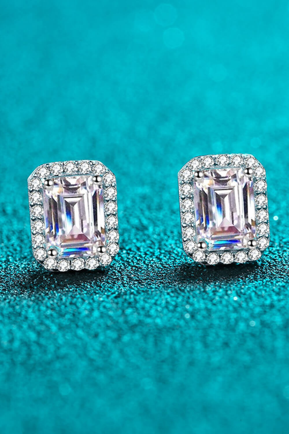 Pair of emerald-cut stud earrings with diamond halo on a teal background