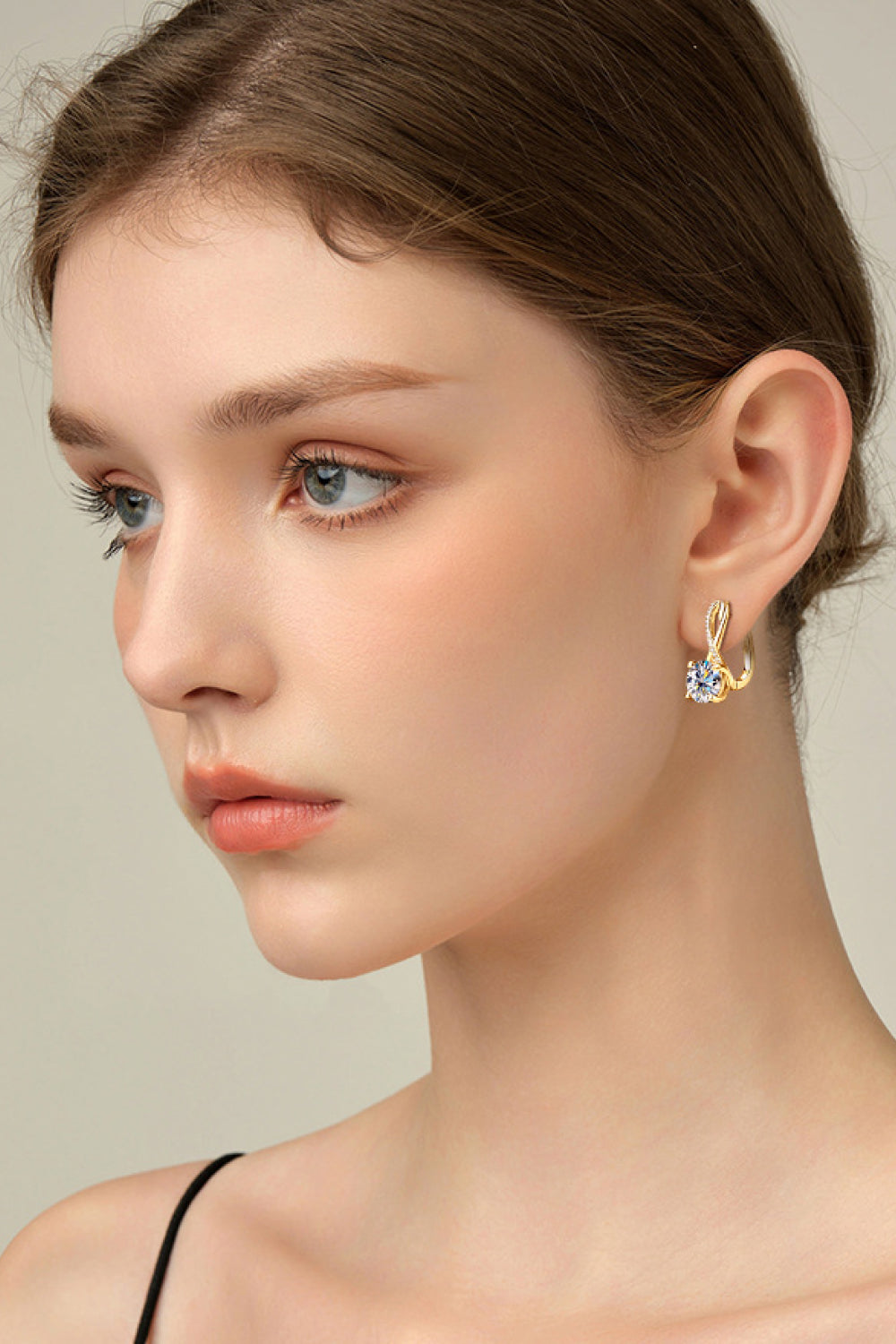 Close-up of a woman wearing a gold earring with a clear gemstone on a neutral background