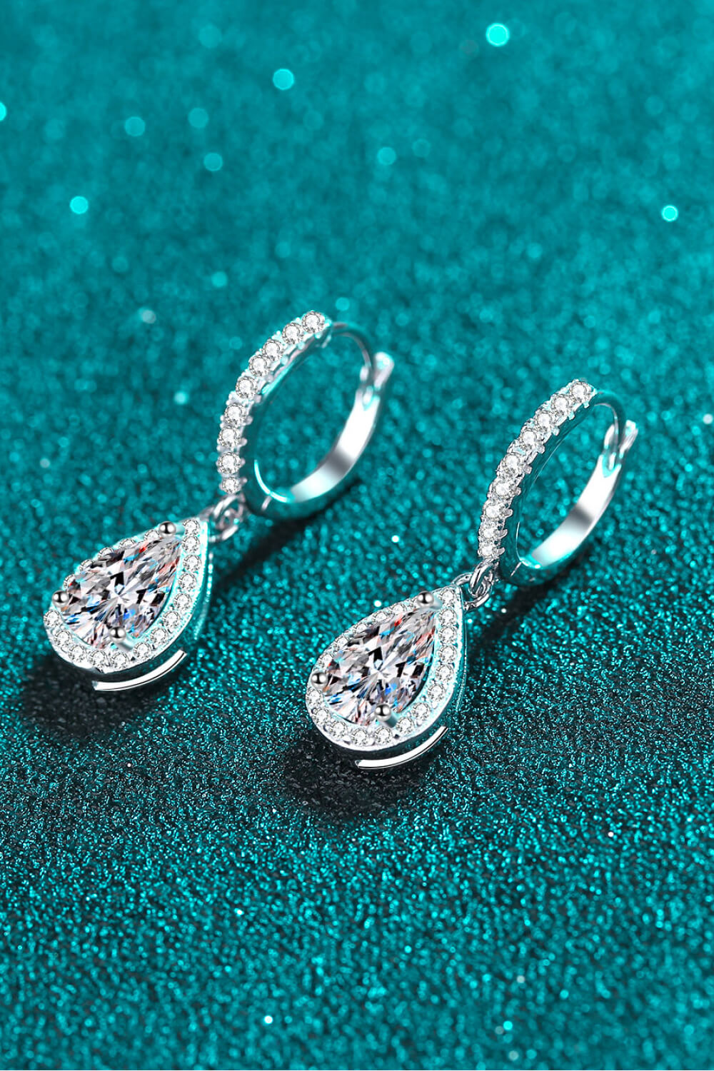 Teardrop-shaped silver earrings with clear gemstones on a teal glittery background